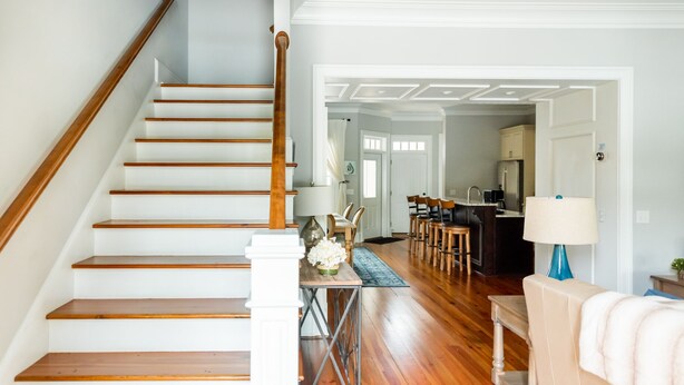 Un escalier avec des marches en bois et une rampe en bois, menant à un espace repas comprenant une table et des chaises, ainsi qu’à une cuisine équipée d’un réfrigérateur et d’un évier.