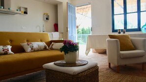 Een woonkamer met een mosterdgele bank, een witte fauteuil en een rieten pouf.