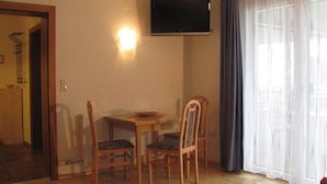 Quarto com mesa de jantar, duas cadeiras, televisão de tela plana na parede e uma janela grande com cortinas leves.