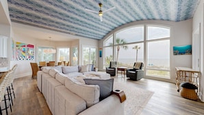 A spacious living room with a large window overlooking a beach, a ceiling with a wave pattern, and a modern chandelier.
