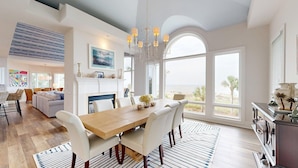 A spacious living room with a large wooden dining table, white chairs, and a fireplace. There is a large window offering a view of the beach.