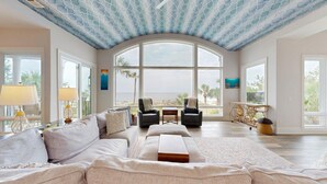 A spacious living room with a large arched window offering a view of the beach, a cozy seating area with a sofa and armchairs, and a decorative ceiling with a wave pattern.