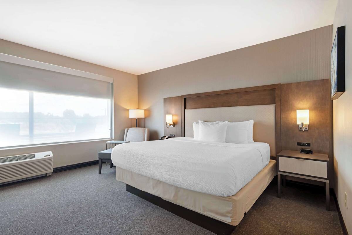 Habitación de hotel con una cama grande, mesitas de noche, una silla y una ventana con persianas.