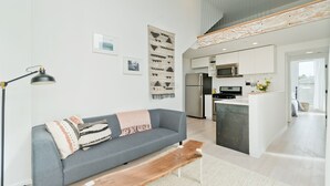 Ein modernes Wohnzimmer mit einem grauen Sofa, einer Holzbank und einer Stehlampe. Im Hintergrund ist eine Küche mit weißen Schränken und einer Mikrowelle zu sehen.