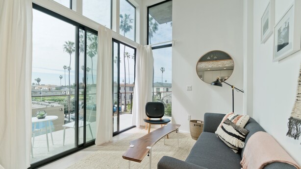 Ein modernes Wohnzimmer mit grauem Sofa, einem Holztisch, einem schwarzen Sessel und einem großen Fenster mit Blick auf Palmen und einen Balkon.