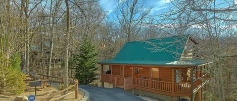 A cabin with a green roof, surrounded by trees, and a gravel driveway leading to it.