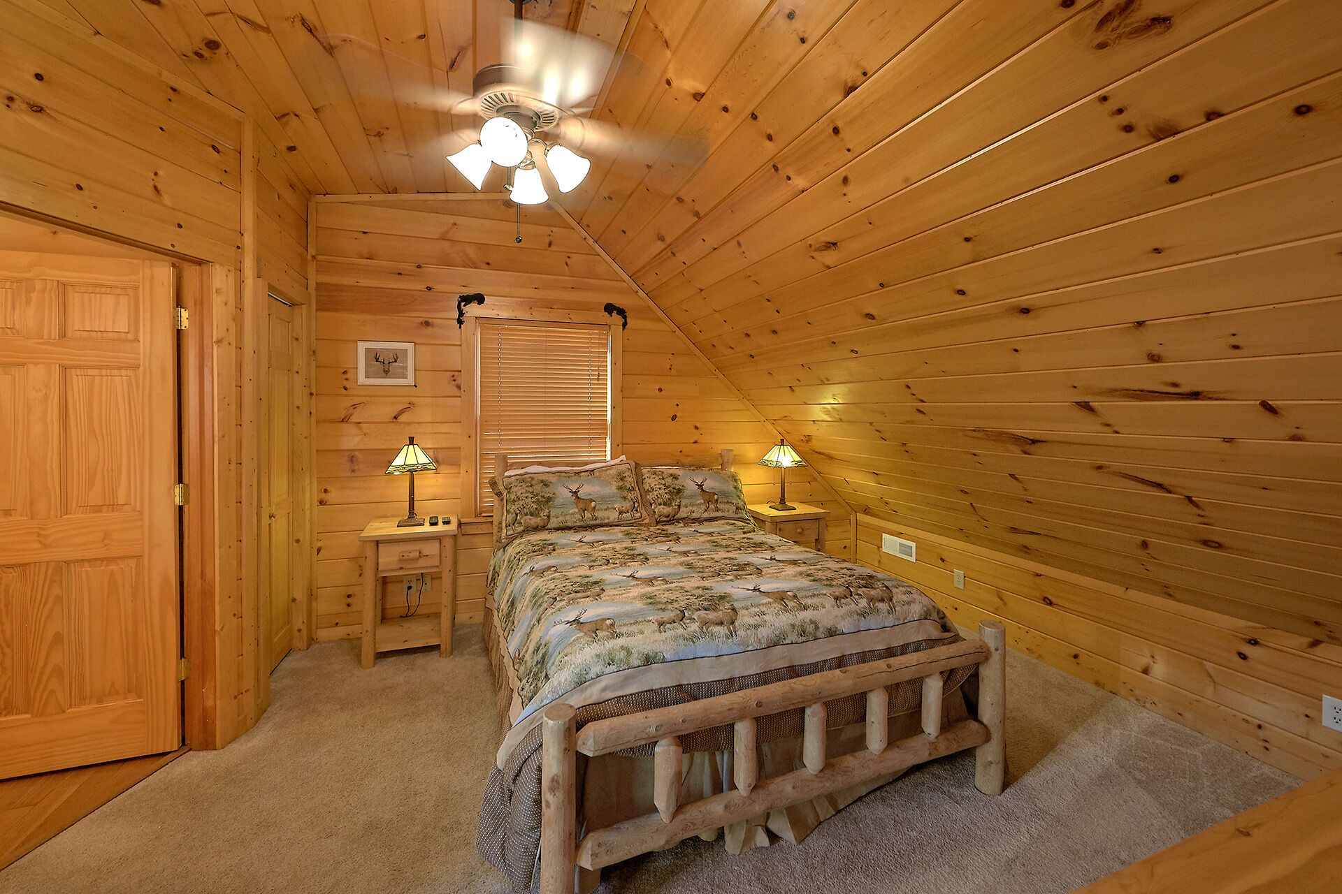 A cozy wooden cabin bedroom with a bed, two bedside tables with lamps, a ceiling fan, and a window with blinds.