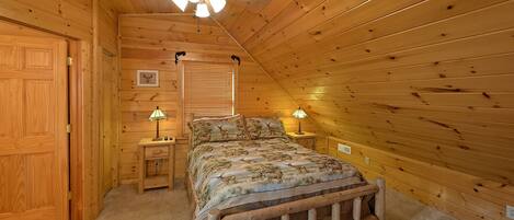 A cozy wooden cabin bedroom with a bed, two bedside tables with lamps, a ceiling fan, and a window with blinds.