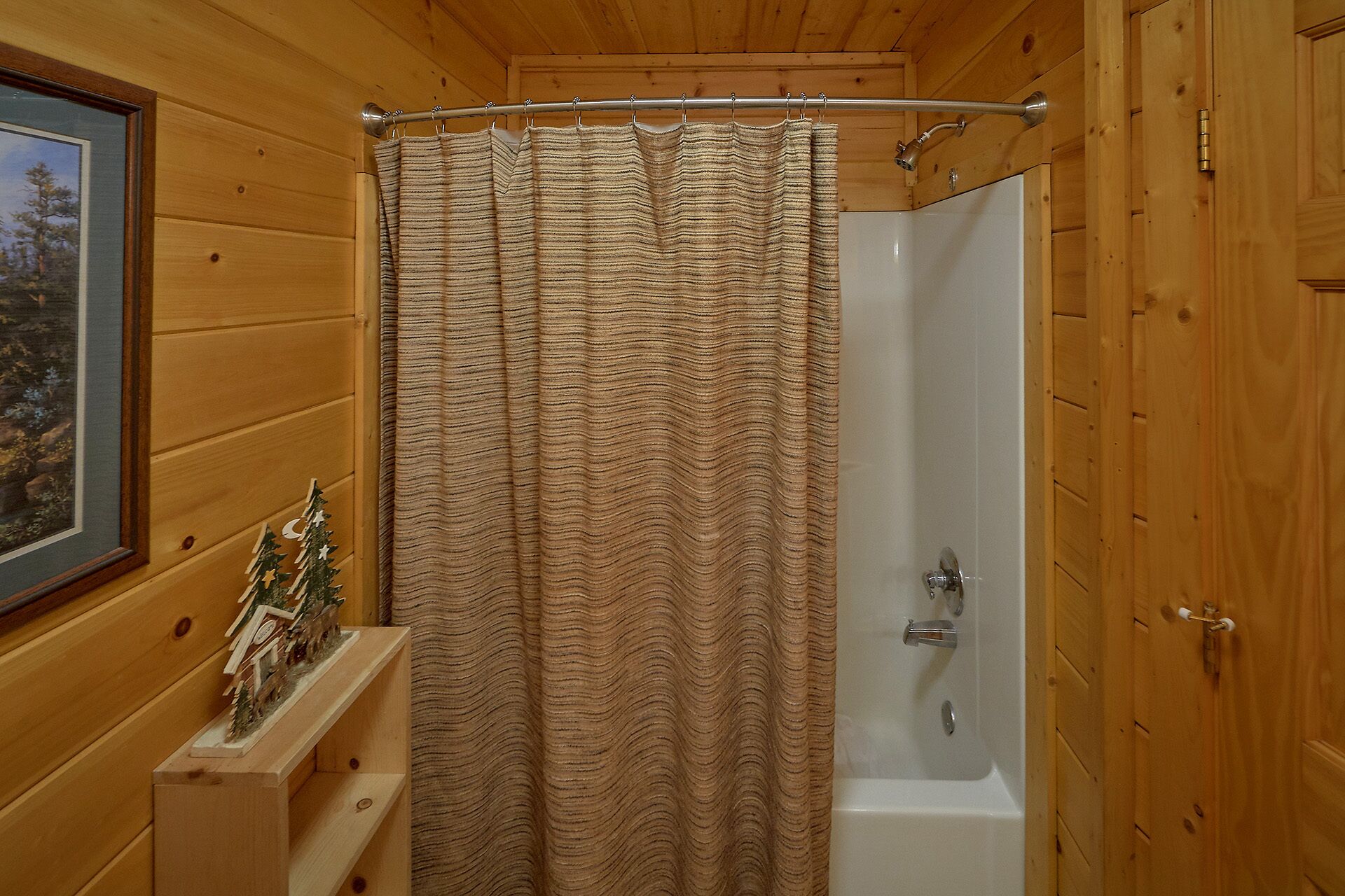 A wooden cabin bathroom with a shower curtain, a small wooden shelf with a Christmas tree decoration, and a framed picture on the wall.