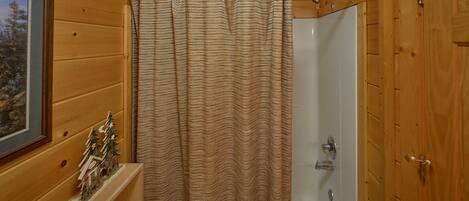 A wooden cabin bathroom with a shower curtain, a small wooden shelf with a Christmas tree decoration, and a framed picture on the wall.