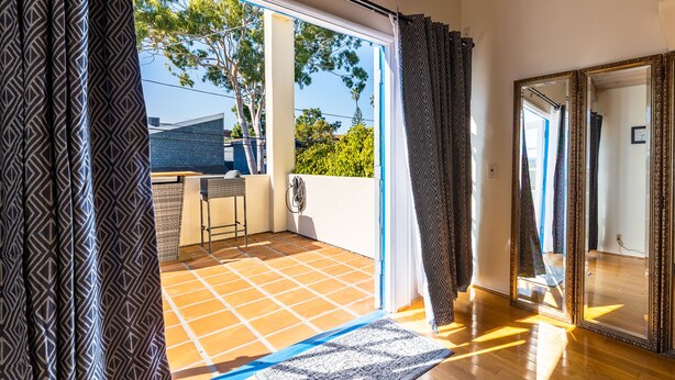 Habitación con un amplio ventanal corredero que da a un balcón con vistas a árboles y un edificio. En el balcón hay una pequeña mesa y una silla, y un espejo grande en la pared.