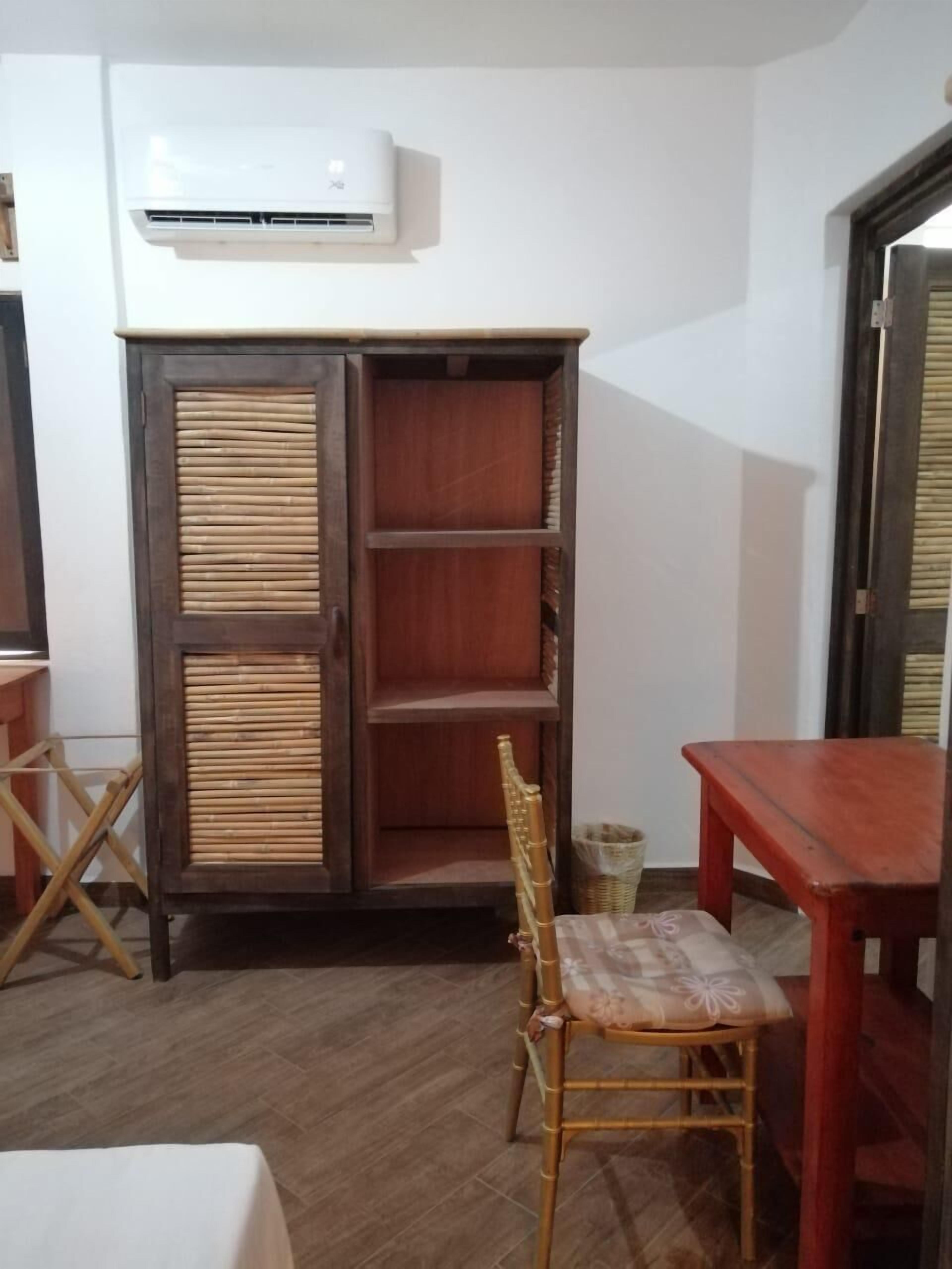 Una habitación con un armario de madera, una silla de mimbre, una mesita pequeña y un equipo de aire acondicionado en la pared.
