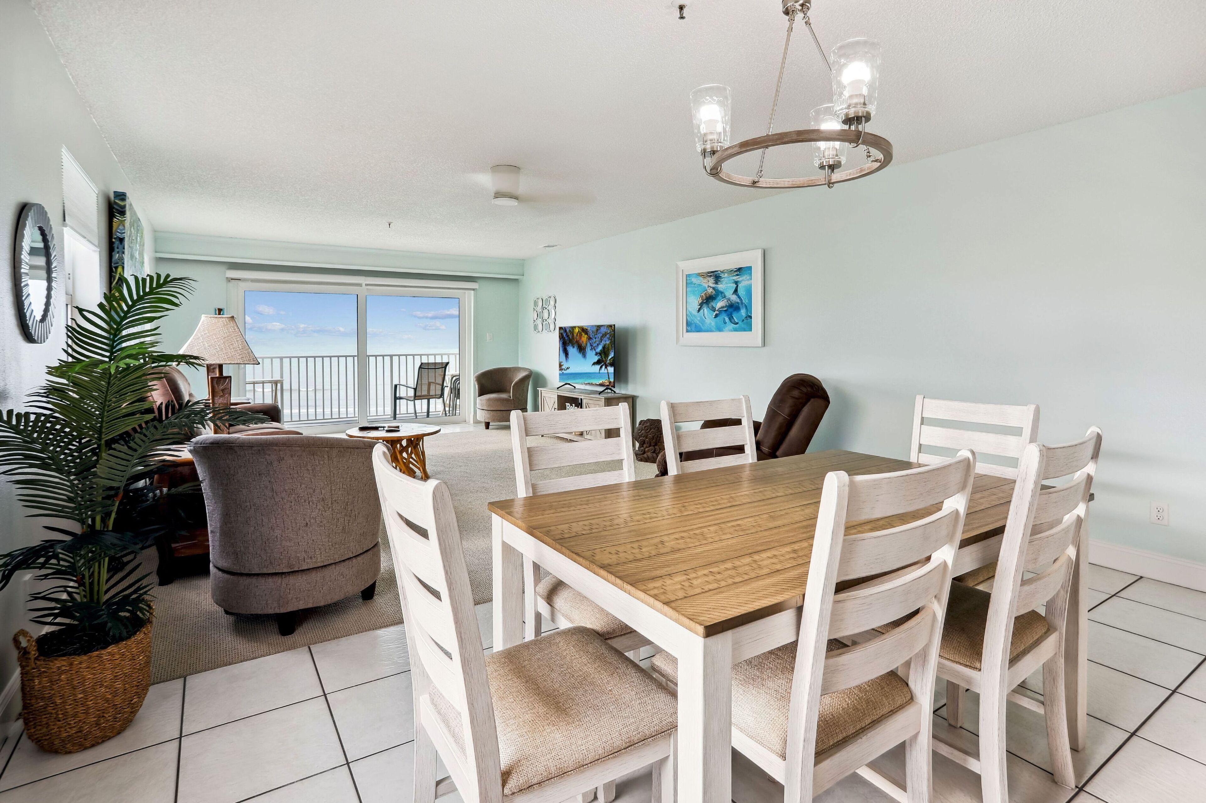A dining area with a wooden table and white chairs, a chandelier, and a view of the ocean through large windows.