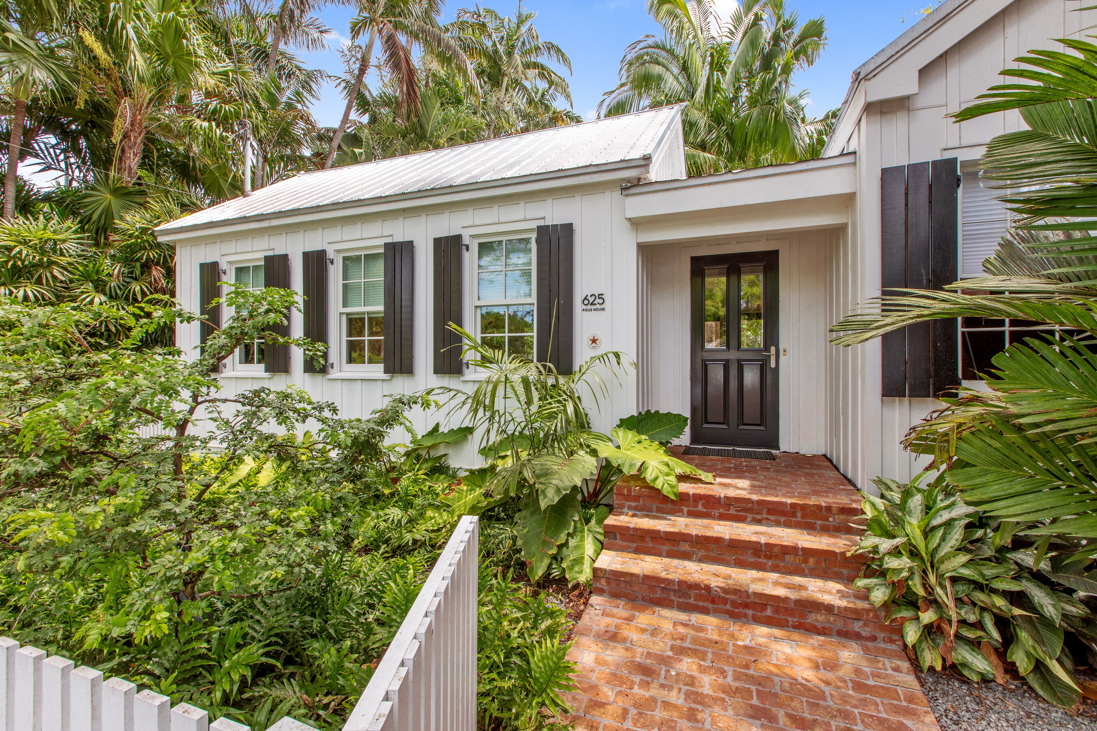 A white building with black shutters, a brick entrance, and lush greenery around it.