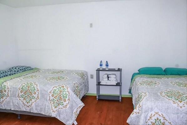 A room with two beds, each with patterned bedding, a small shelf with folded towels, and a water bottle on the shelf.