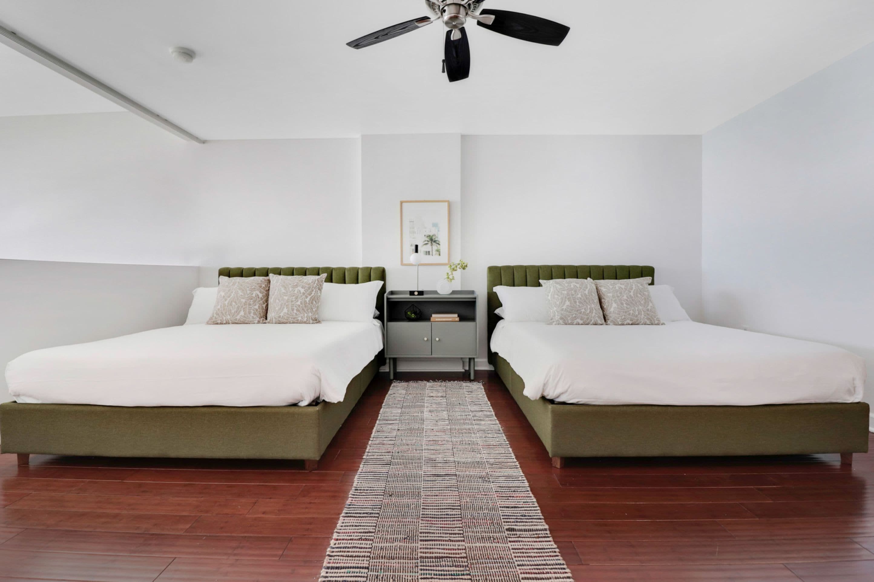 A hotel room with two beds, a ceiling fan, a nightstand, and a framed picture on the wall.