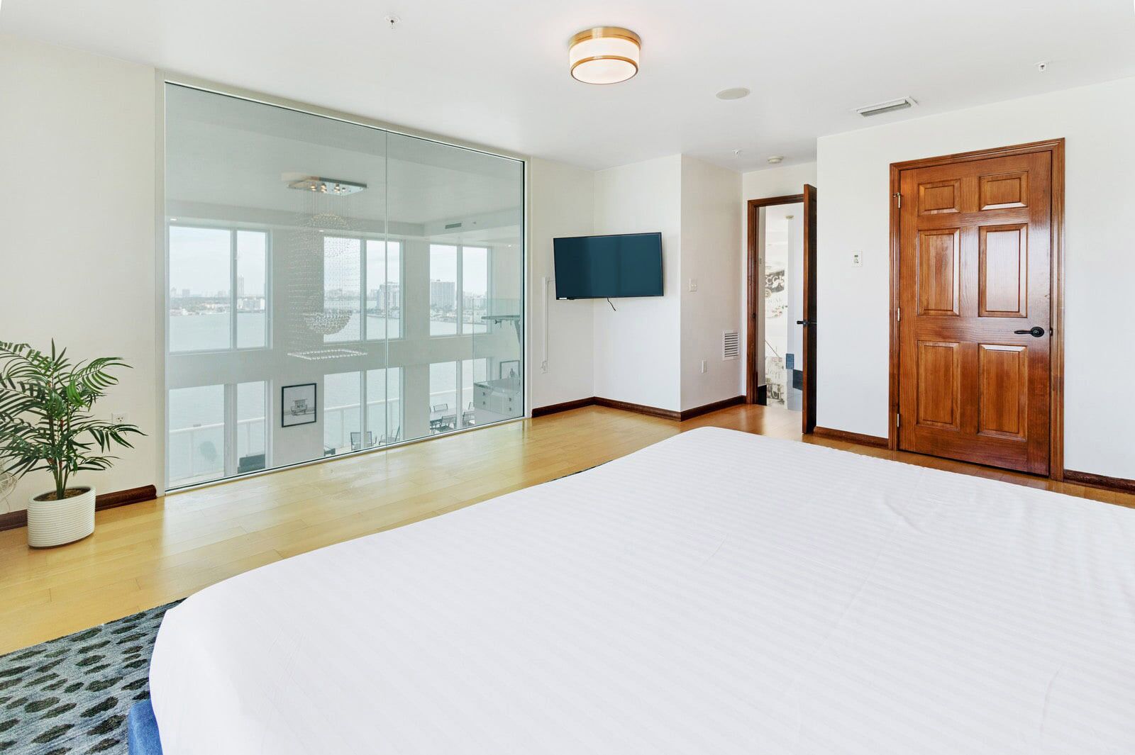 A modern bedroom with a large bed, a glass wall with a city view, a wooden door, and a mounted TV.