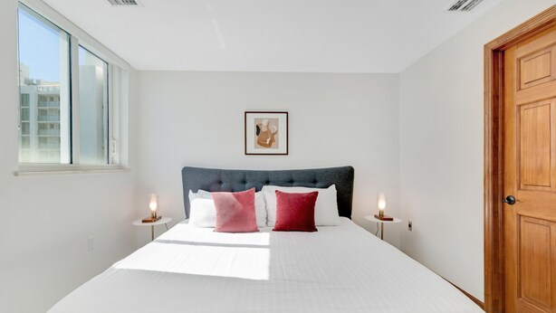 A bedroom with a large bed, two bedside tables, a wooden door, and a window. - Roami at Adagio (North Bay Village)