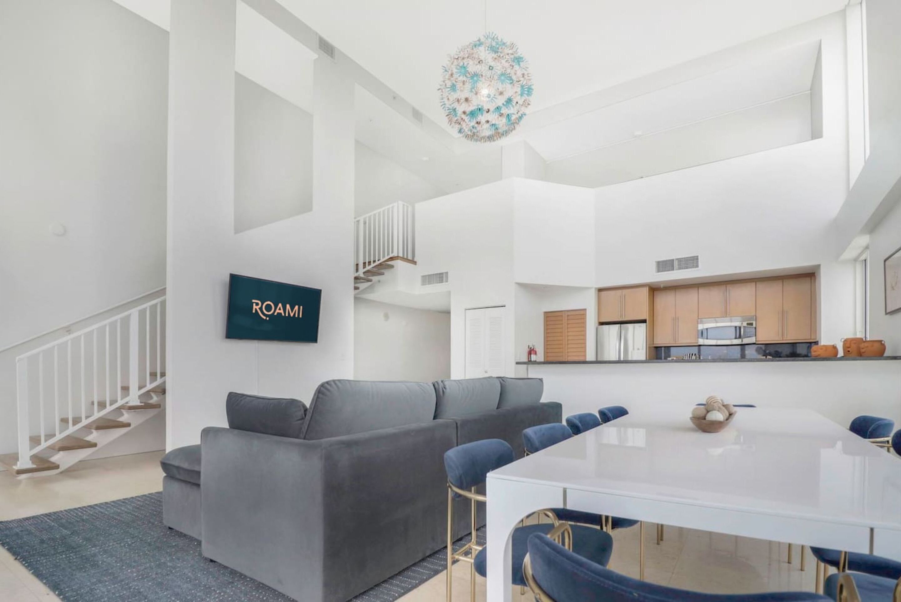 Ein modernes Wohnzimmer mit grauem Sofa, einem Esstisch mit blauen Stühlen und einer Treppe, die in eine obere Ebene führt.