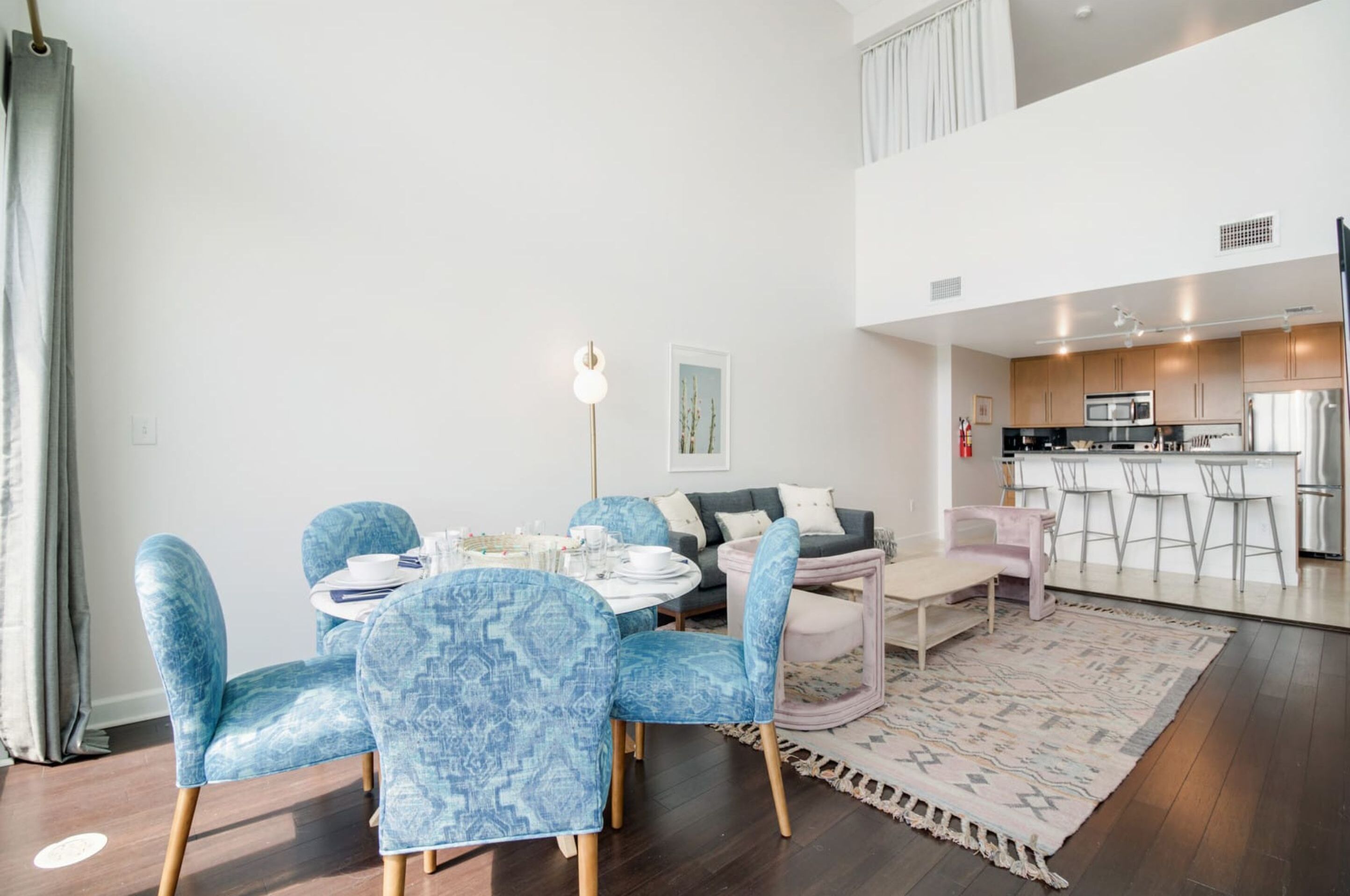 A modern living room with a dining area, featuring blue patterned chairs, a round table, and a grey sofa. A kitchen is visible in the background with stainless steel appliances.