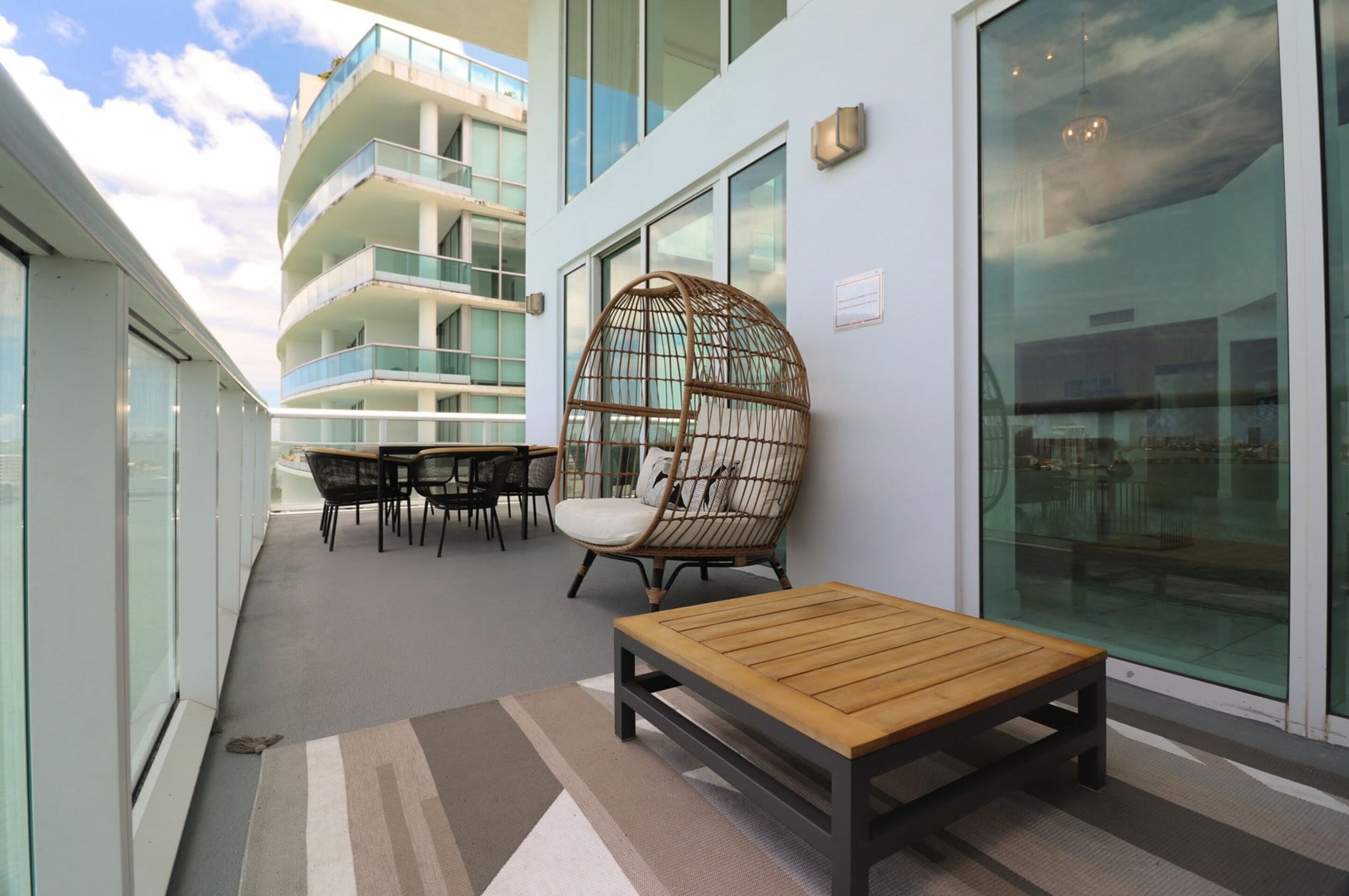A modern balcony with a wooden table, wicker chairs, and a glass railing overlooking a building.