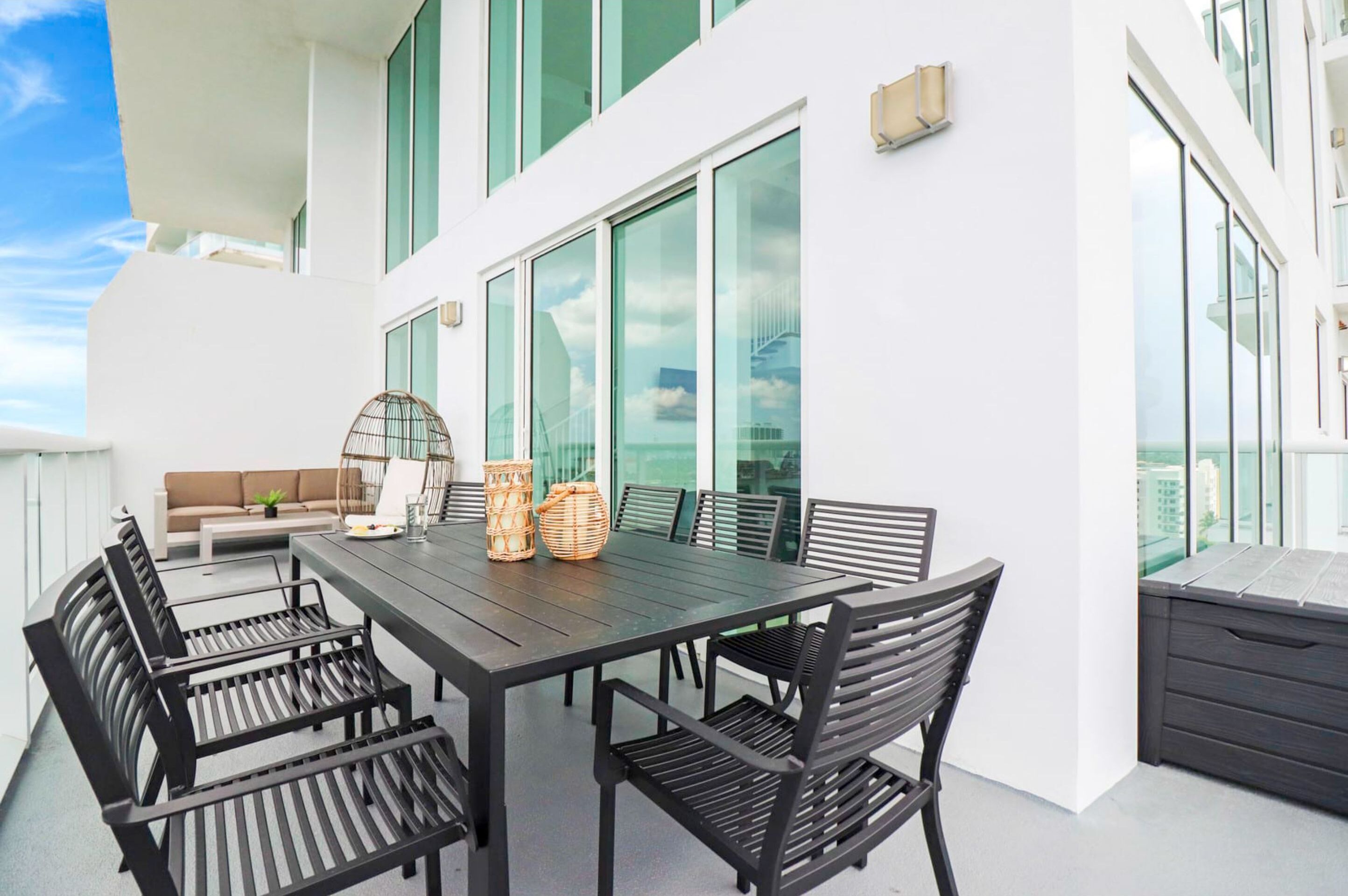 A modern outdoor dining area with a dark wooden table and black metal chairs, surrounded by large windows and a white wall.