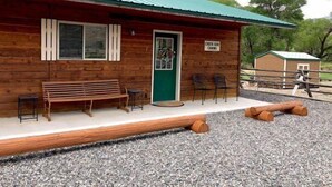 Une cabane en bois avec une porte verte, un banc et deux chaises à l’extérieur.