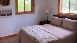 Une chambre à coucher avec un lit, deux fenêtres, une table de chevet avec une lampe et des œuvres d’art murales.