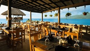 A restaurant with wooden tables and chairs, a thatched roof, and hanging plants.