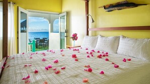 A room with a bed covered in rose petals, a view of the sea through an open door, and a wooden headboard.