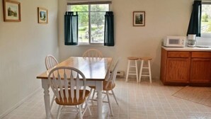 Un coin repas avec une table en bois et des chaises blanches, une fenêtre avec des rideaux, et un espace cuisine équipé de placards et d’un four à micro-ondes.