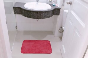 Un bagno con lavandino bianco, piano di lavoro in granito scuro, pareti piastrellate di bianco, tappetino da bagno rosso e water bianco.