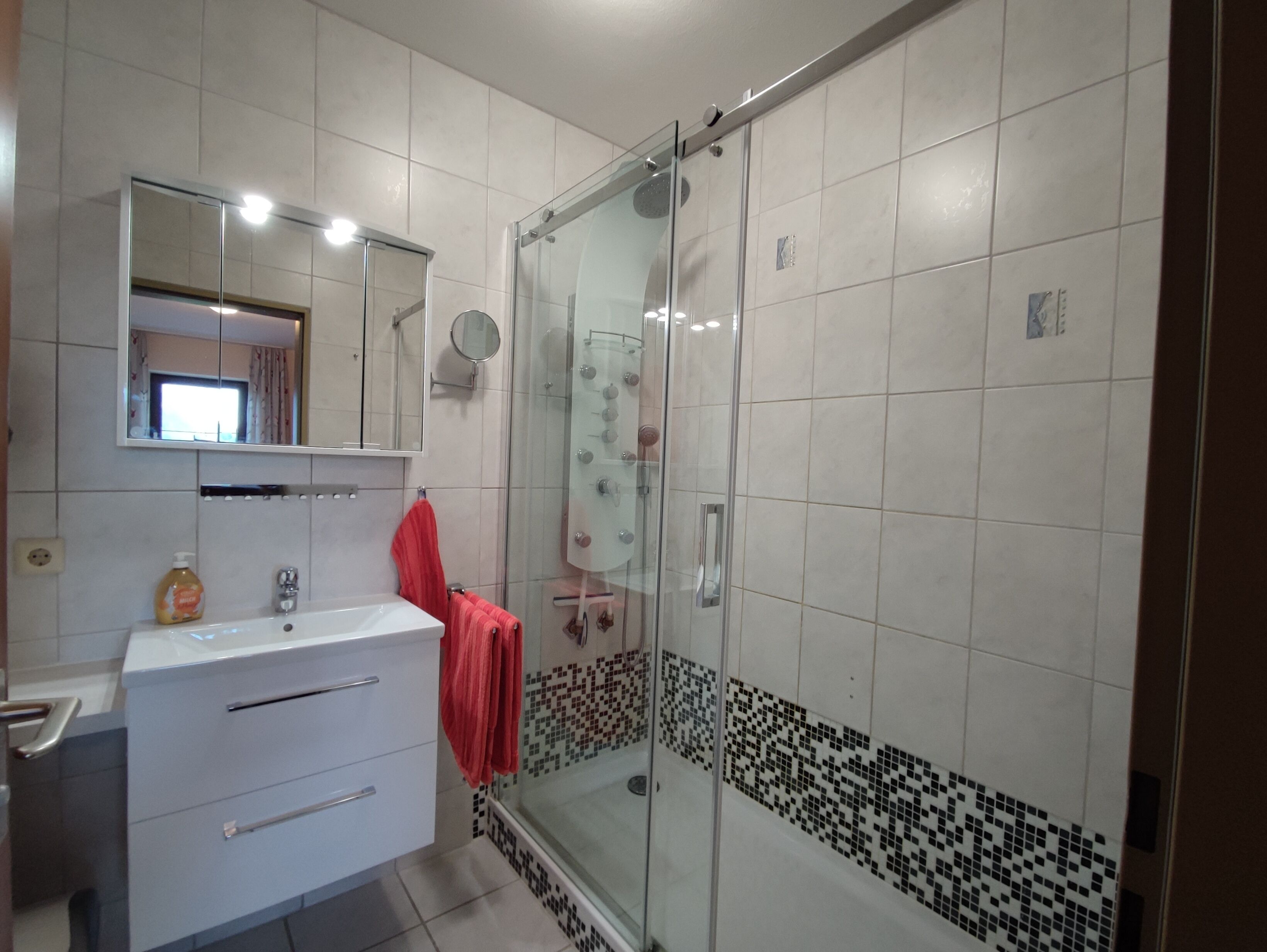 Casa de banho com cabine de duche em vidro, lavatório branco com espelho em cima e toalha vermelha pendurada na parede.