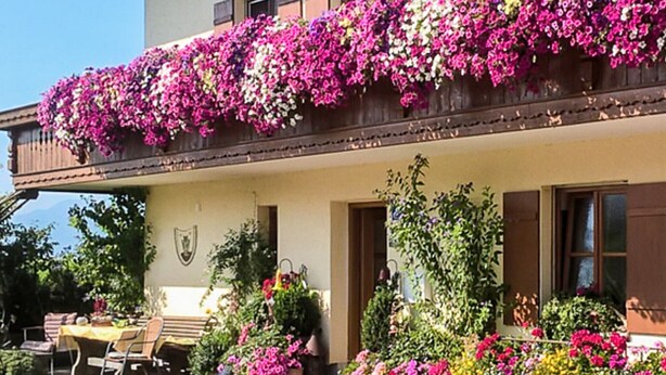 A building with a balcony covered in colorful flowers, a dining area with outdoor seating, and a garden with various plants. - Vacation apartment with south-facing balcony (Riedering)