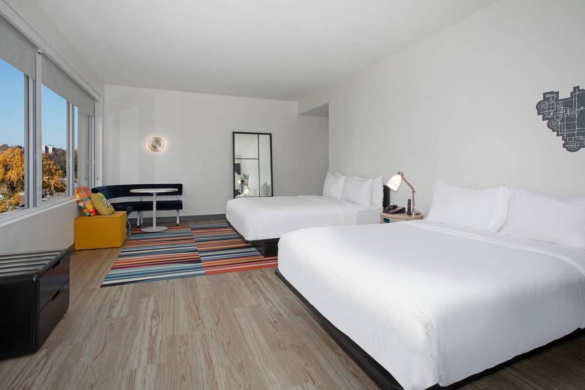 A modern hotel room with two beds, a desk, a chair, and a large window.