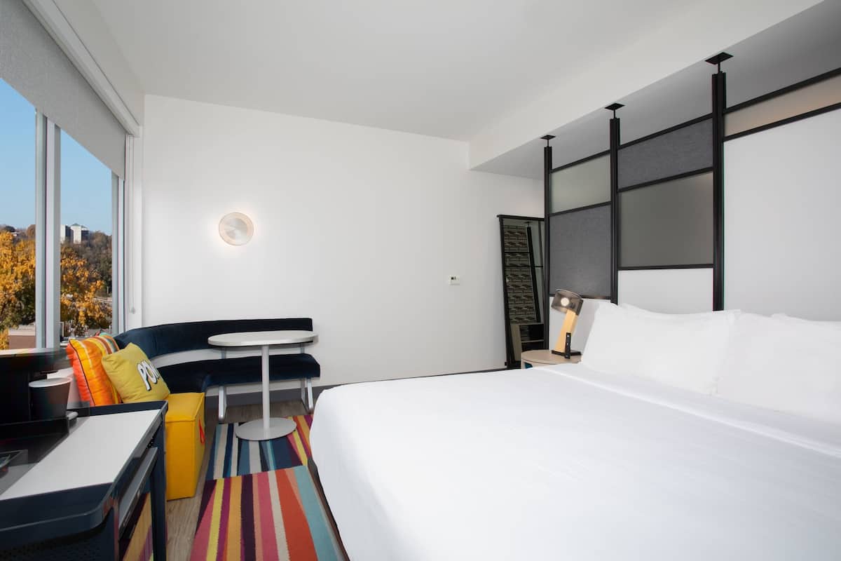 A modern hotel room with a large bed, a small round table, a sofa with colorful cushions, and a view of the outdoors through large windows.