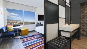 A modern hotel room with a large window, a bed, a desk, a bathroom, and a colorful striped rug. - Aloft by Marriott Omaha Aksarben Village (Omaha)