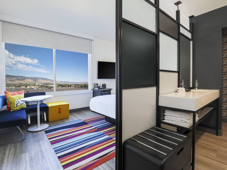 Une chambre d’hôtel moderne dotée d’une grande fenêtre, d’un lit, d’un bureau, d’une salle de bain et d’un tapis rayé aux couleurs vives.