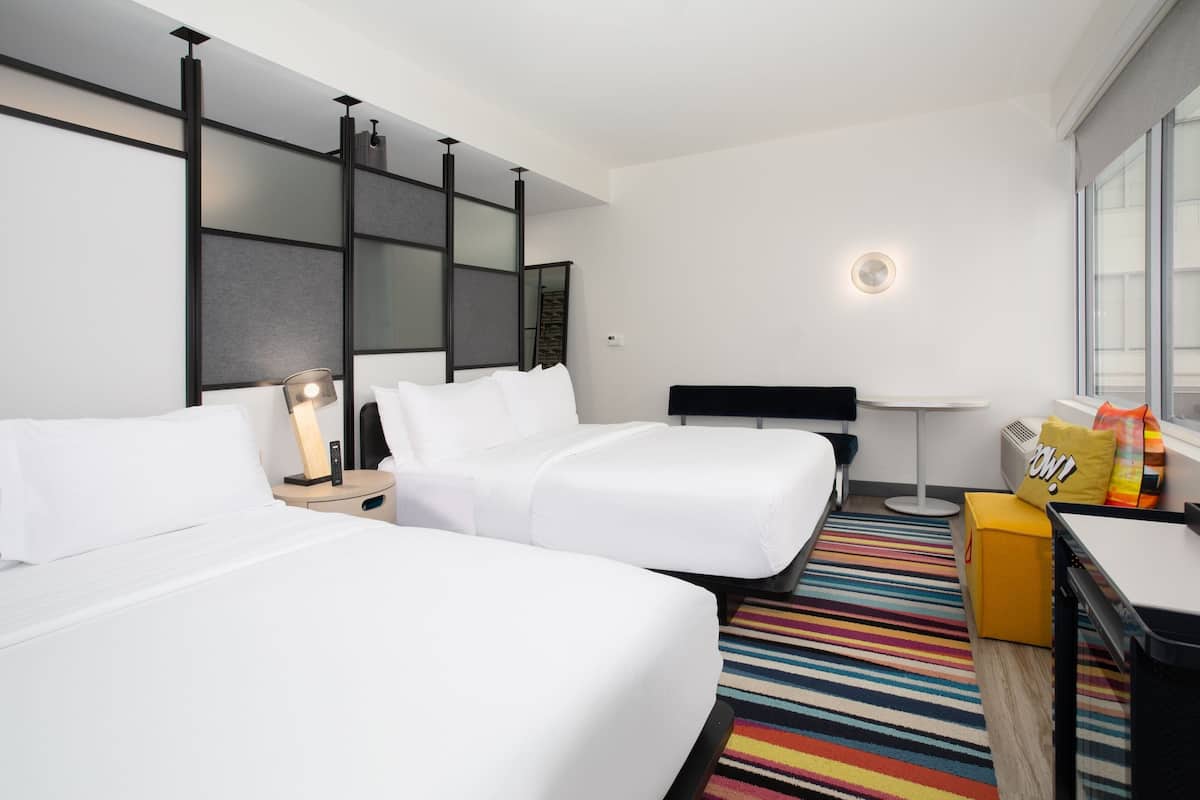 A hotel room with two beds, a desk, a chair, a colorful striped rug, and a window with a view.