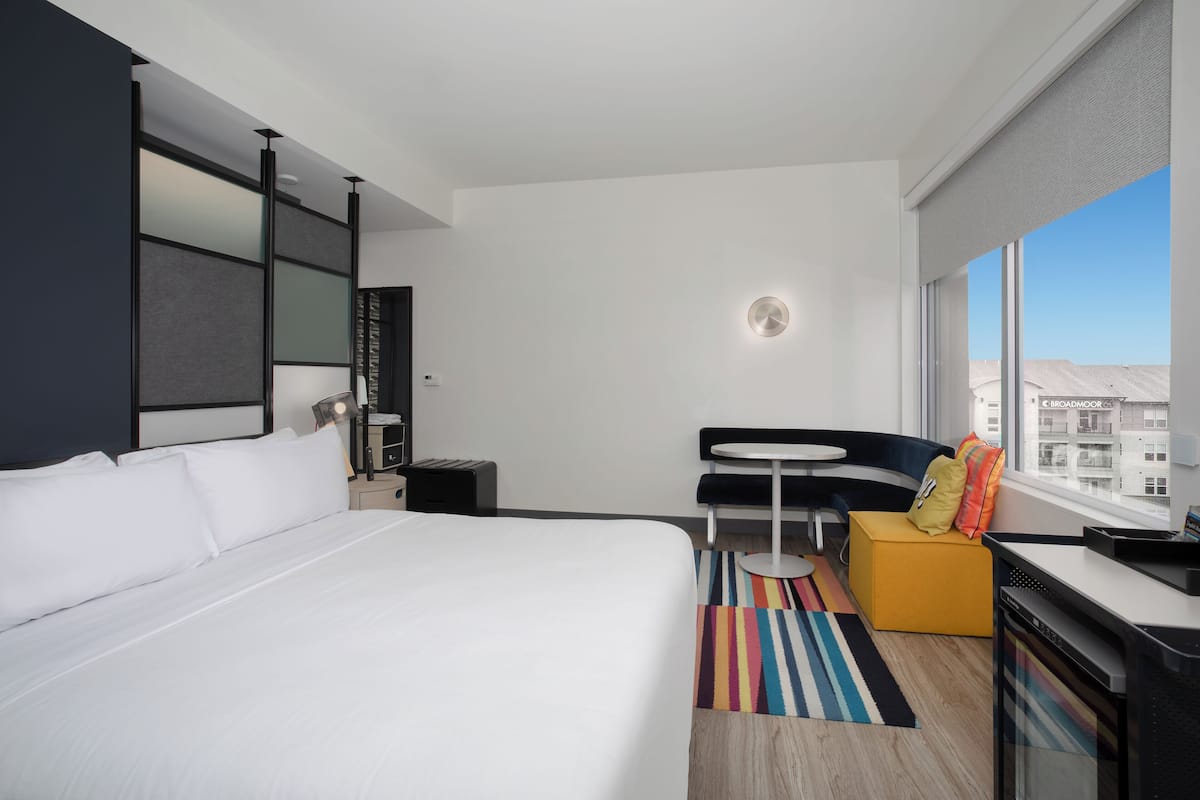 A modern hotel room with a large bed, a small desk, a chair, a sofa, and a window with a city view.