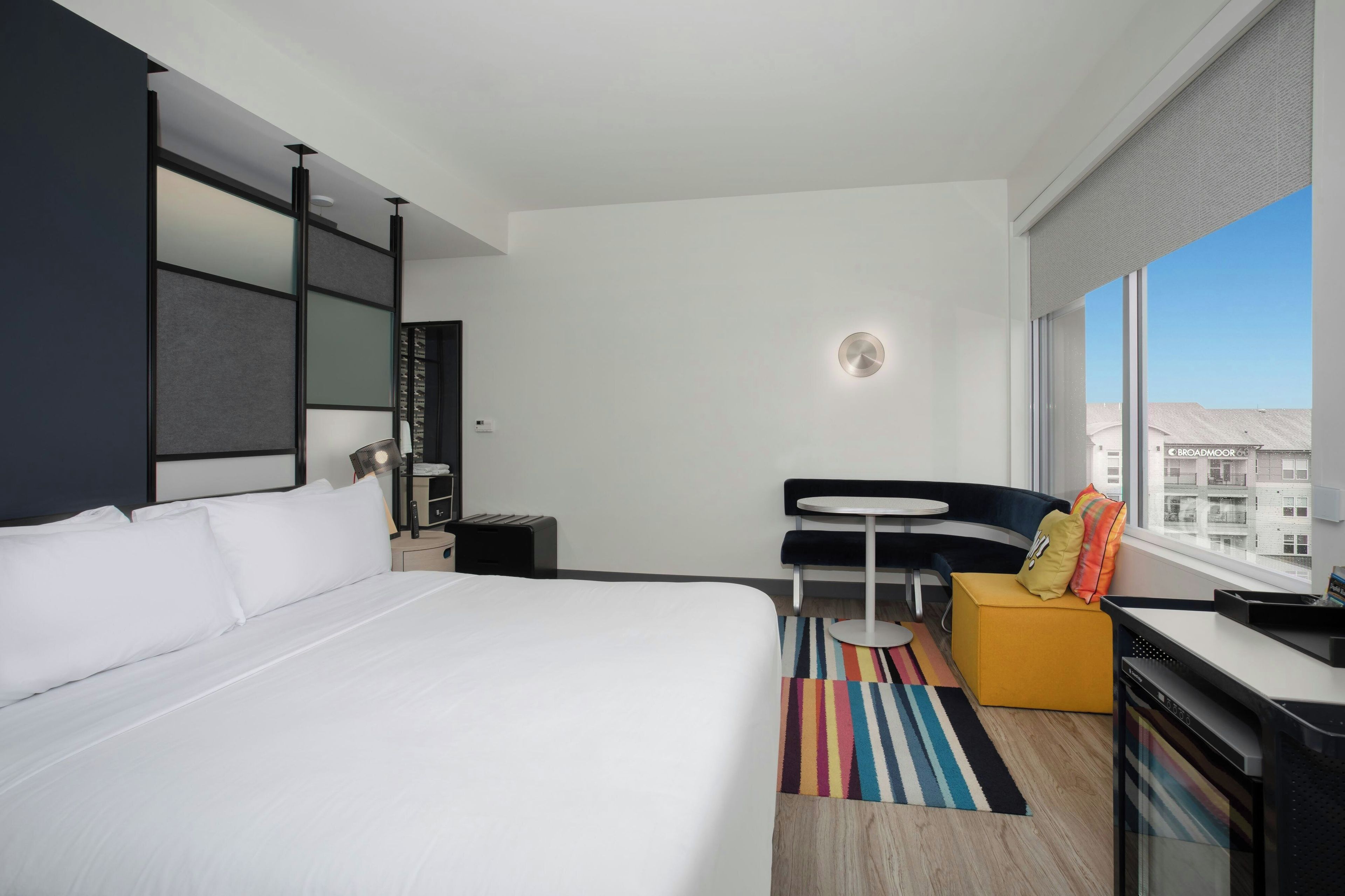 Een hotelkamer met een groot bed, een klein bureau, een stoel, een ronde tafel, een kleurrijk gestreept tapijt en een raam met uitzicht op de stad.