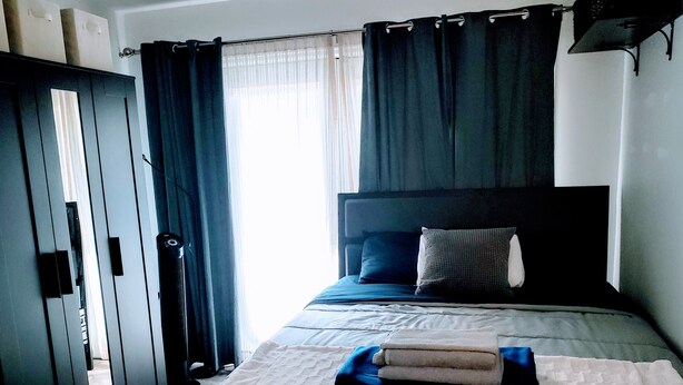 Ein ordentlich bezogenes Bett mit weißer Decke und gefaltetem Handtuch, ein dunkles Kopfteil und ein Fenster mit durchsichtigen Vorhängen.
