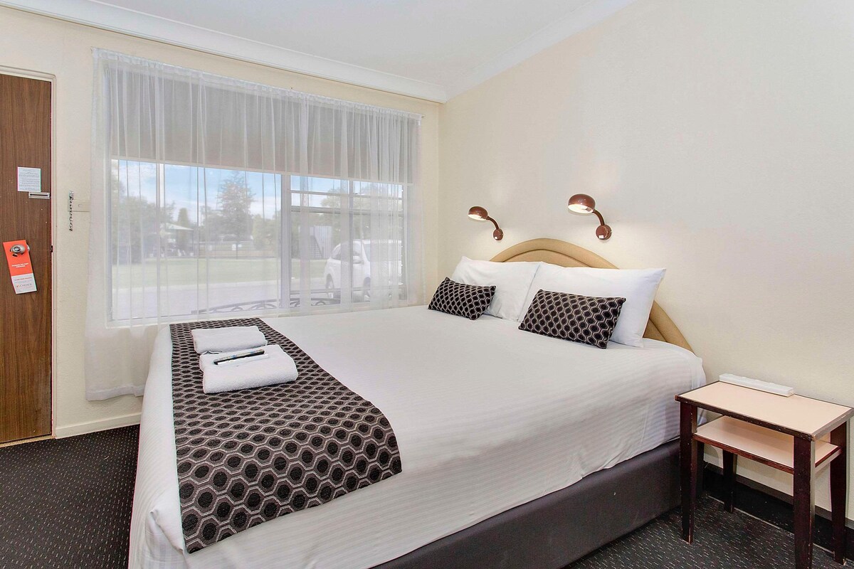 A hotel room with a large bed, a bedside table, a window with curtains, and a door with a sign.