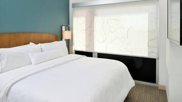 Quarto de hotel com uma cama grande, mesa de cabeceira com abajur e janela com desenho ondulado.