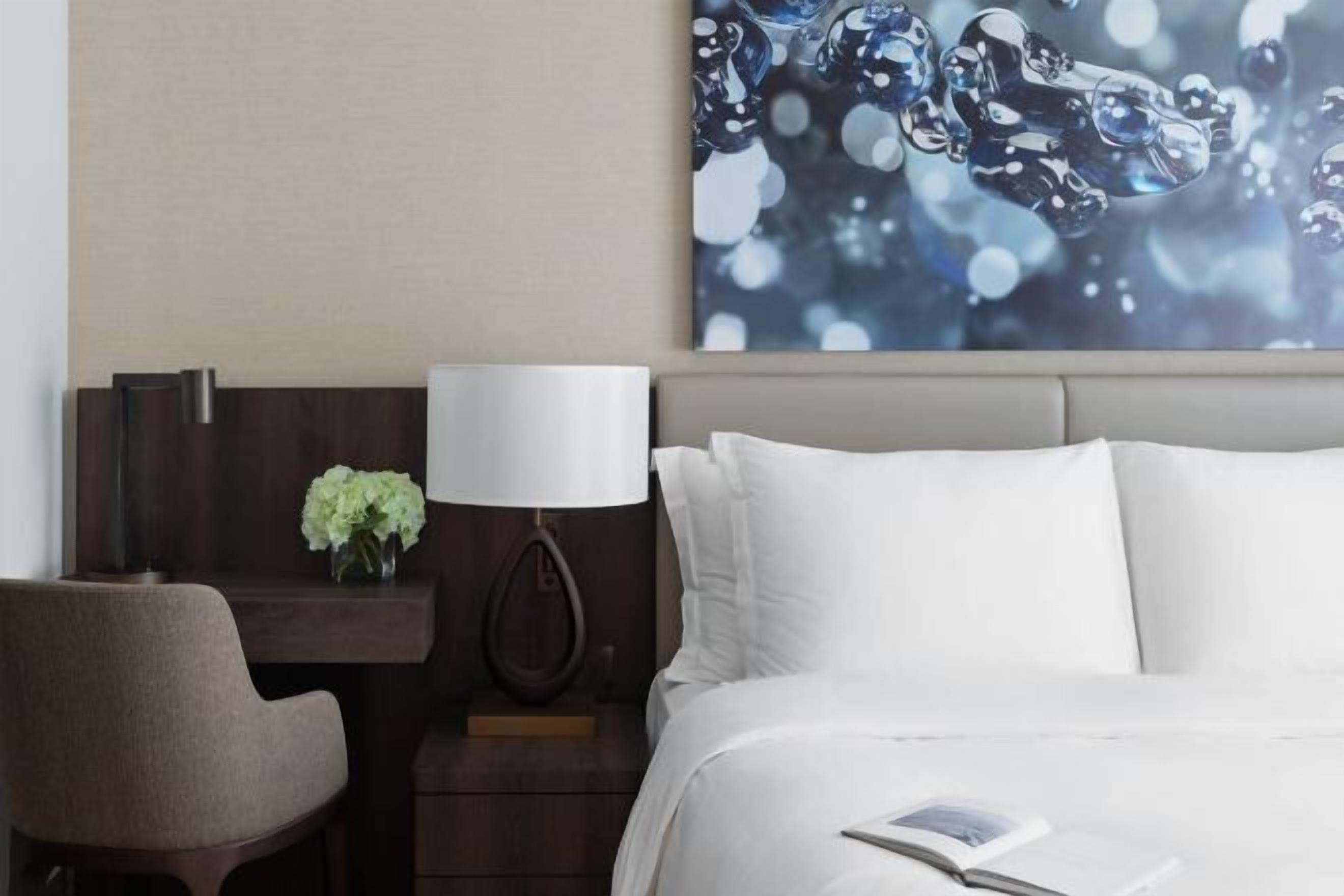 A hotel room with a bed, a desk with a lamp, and a wall-mounted artwork.