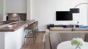 A modern kitchen with a central island, a dining area with a round table, and a living space with a sofa and a flat-screen TV mounted on the wall.