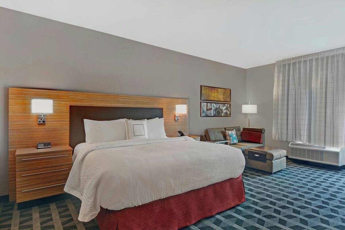 A hotel room with a large bed, a wooden headboard, a bedside table with a lamp, a small sofa, and a window with curtains.