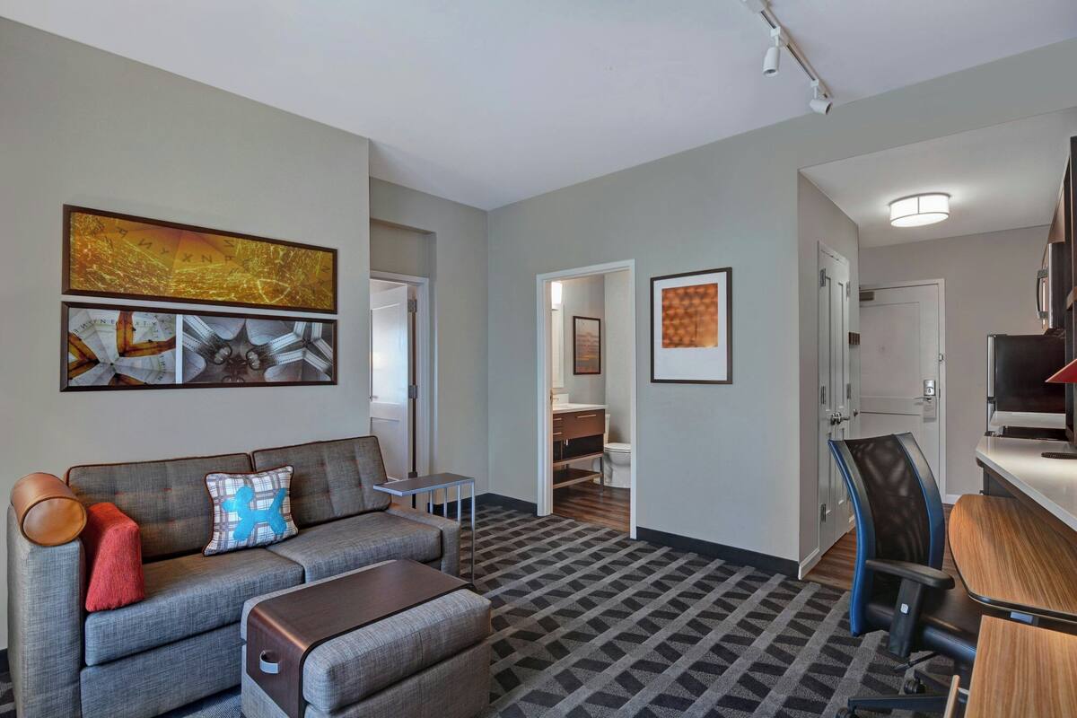 A modern living room with a grey sofa, a small table, and a TV. There is a hallway leading to a bathroom and a bedroom.