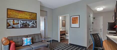 Una sala moderna con un sofá gris, una mesita y un televisor. Se ve un pasillo que conduce a un baño y un dormitorio.