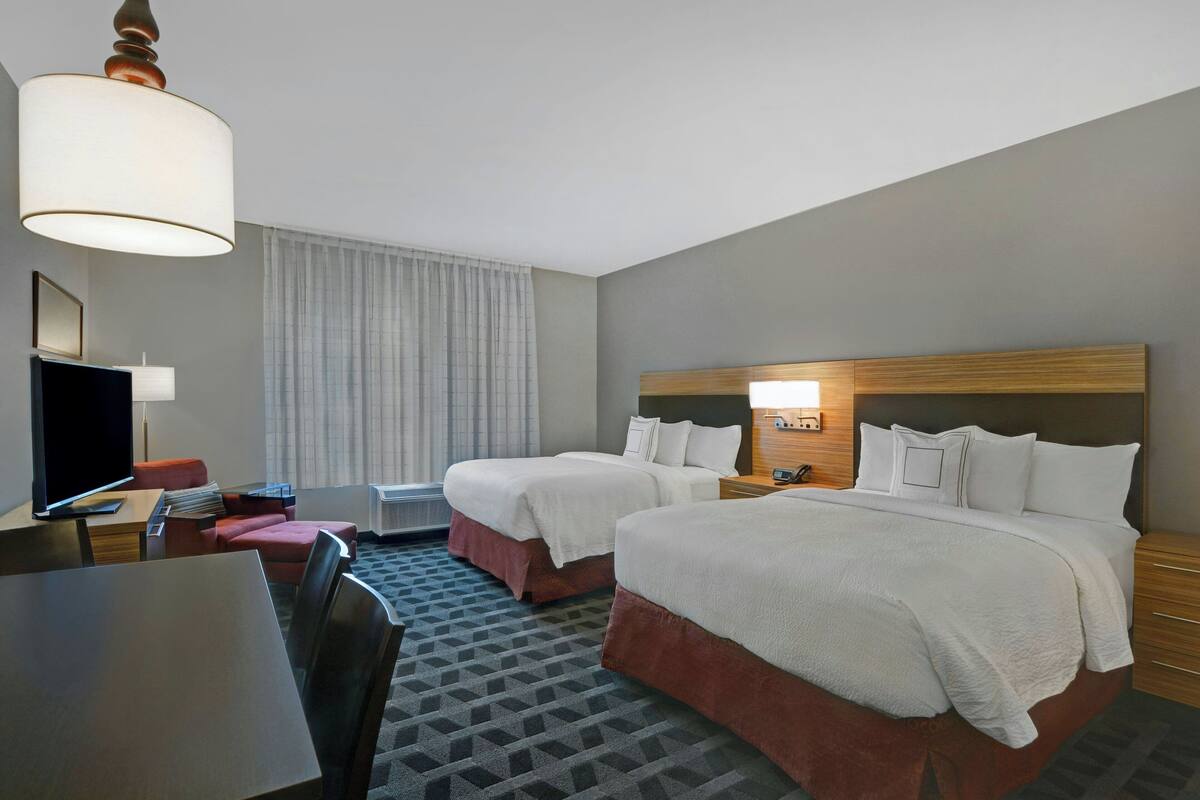 A hotel room with two beds, a desk, a chair, a TV, and a lamp.